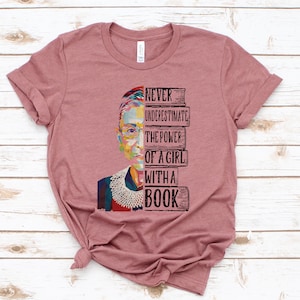 Op de afbeelding: Een mauvekleurig T-shirt met een kleurrijke afbeelding van een vrouwengezicht en de tekst "NEVER UNDERESTIMATE THE POWER OF A GIRL WITH A BOOK". Het shirt is gemaakt van zacht materiaal.