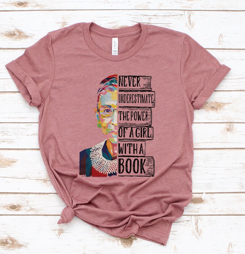 Op de afbeelding: Een mauvekleurig T-shirt met een kleurrijke afbeelding van een vrouwengezicht en de tekst "NEVER UNDERESTIMATE THE POWER OF A GIRL WITH A BOOK". Het shirt is gemaakt van zacht materiaal.