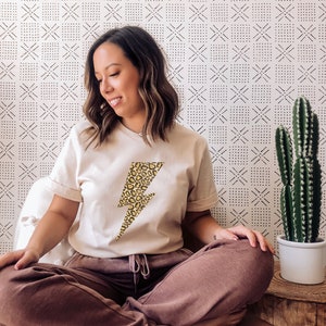 May include: A woman wearing a beige t-shirt with a yellow and brown leopard print lightning bolt graphic. She is sitting on a brown leather chair with her legs crossed. The background is a white wall with a black and white geometric pattern.