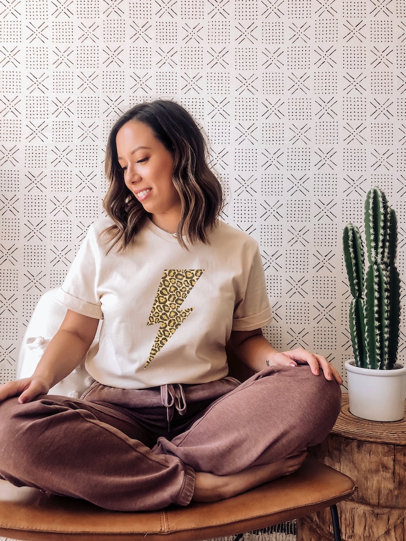 May include: A woman wearing a beige t-shirt with a yellow and brown leopard print lightning bolt graphic. She is sitting on a brown leather chair with her legs crossed. The background is a white wall with a black and white geometric pattern.