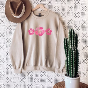 May include: A beige sweatshirt with a pink hibiscus flower design. The sweatshirt is hanging on a hanger against a white background with a black geometric pattern.