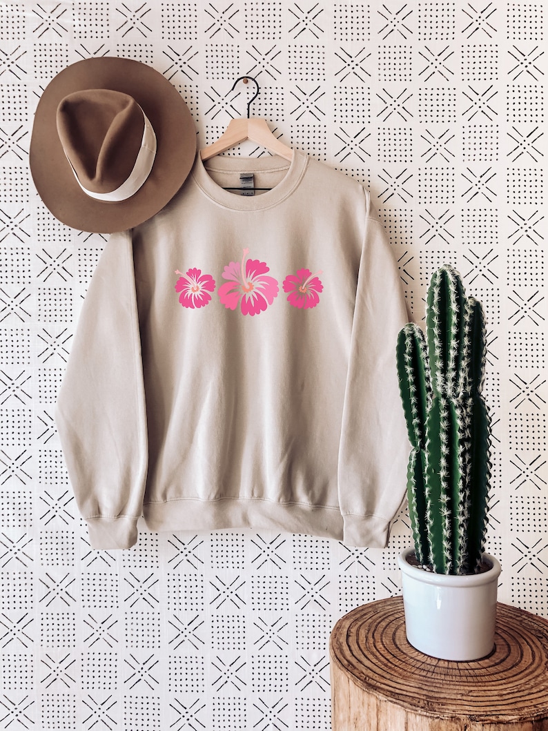 May include: A beige sweatshirt with a pink hibiscus flower design. The sweatshirt is hanging on a hanger against a white background with a black geometric pattern.