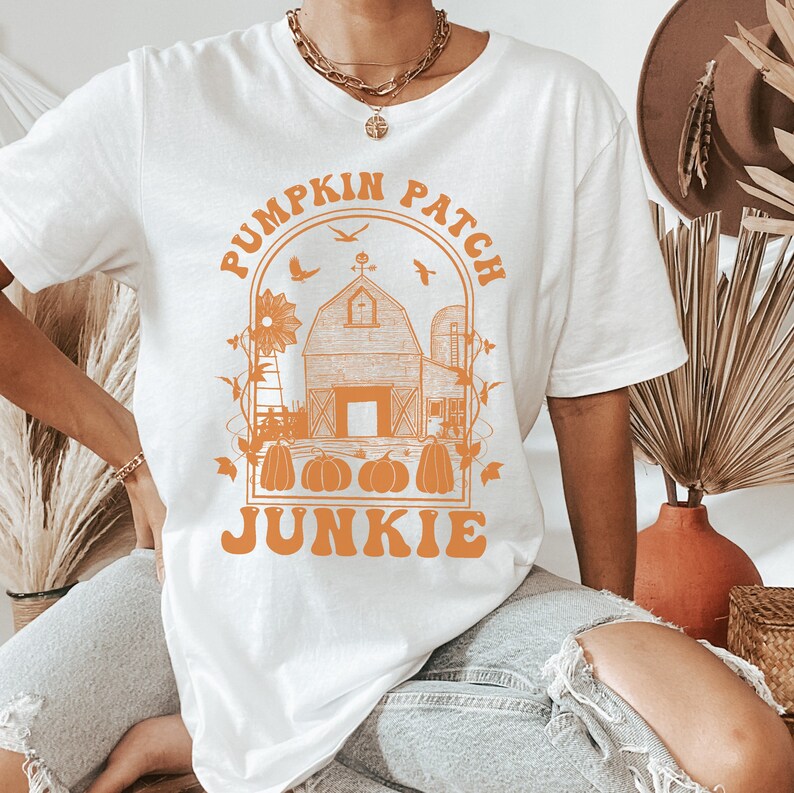 Puede incluir: Camiseta blanca con un gr&aacute;fico naranja de una granja, un molino de viento y calabazas. El texto "Pumpkin Patch Junkie" est&aacute; escrito en una fuente retro.