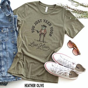 Puede incluir: Camiseta verde con una rana de dibujos animados que lleva un sombrero de vaquero y sostiene una pistola. El texto de la camiseta dice "You just yeed your last haw".