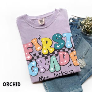 May include: A light purple t-shirt with the words "FIRST GRADE" in colorful bubble letters, a black and white checkered pattern, and the name "Mrs. Jackson". The shirt is paired with blue jeans.