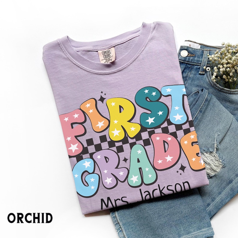 May include: A light purple t-shirt with the words "FIRST GRADE" in colorful bubble letters, a black and white checkered pattern, and the name "Mrs. Jackson". The shirt is paired with blue jeans.