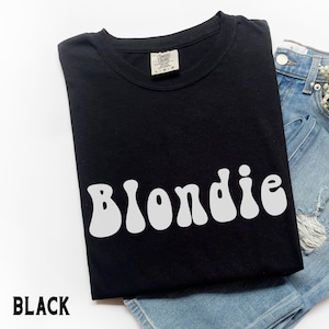 Puede incluir: Camiseta negra con la palabra "Blondie" en blanco, con letras de estilo retro. La camiseta est&aacute; doblada y colocada sobre unos pantalones cortos vaqueros azules. La palabra "BLACK" est&aacute; impresa en blanco en la parte inferior izquierda.