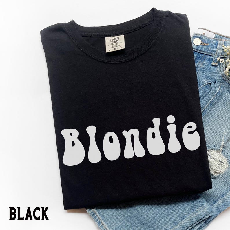 Puede incluir: Camiseta negra con la palabra "Blondie" en blanco, con letras de estilo retro. La camiseta est&aacute; doblada y colocada sobre unos pantalones cortos vaqueros azules. La palabra "BLACK" est&aacute; impresa en blanco en la parte inferior izquierda.