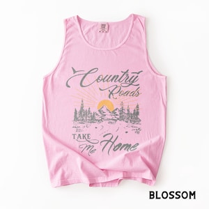 K&ouml;nnte beinhalten: Ein hellrosa Tanktop mit einem Aufdruck, der "Country Roads Take Me Home" in grauer Schrift zeigt. Die Grafik zeigt eine Berglandschaft mit aufgehender Sonne. Das Wort "Blossom" ist unten aufgedruckt.