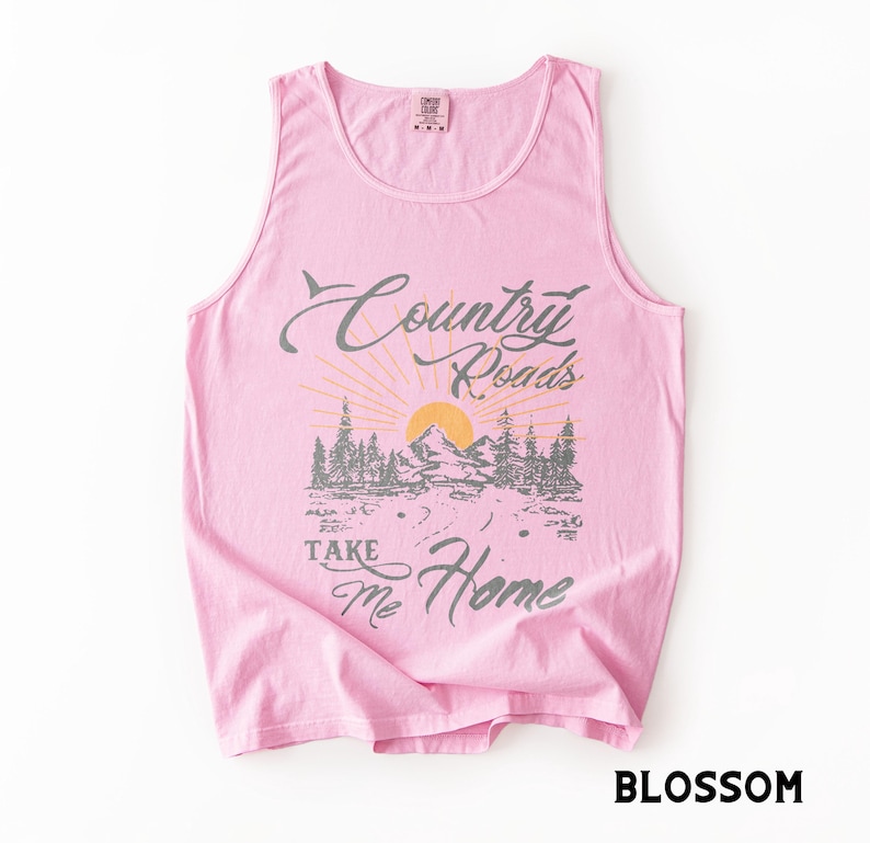 K&ouml;nnte beinhalten: Ein hellrosa Tanktop mit einem Aufdruck, der "Country Roads Take Me Home" in grauer Schrift zeigt. Die Grafik zeigt eine Berglandschaft mit aufgehender Sonne. Das Wort "Blossom" ist unten aufgedruckt.