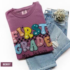 May include: A berry-colored t-shirt with the words "FIRST GRADE" in colorful bubble letters, with stars and a checkered pattern. The text "Mrs. Jackson" is below the main text. The shirt is paired with light blue denim jeans.