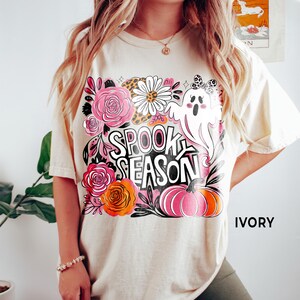May include: Ivory-colored t-shirt with a colorful "Spooky Season" graphic. The design includes a ghost, pumpkins, flowers, and the text "Spooky Season". The shirt is a relaxed fit.