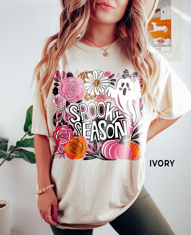 May include: Ivory-colored t-shirt with a colorful "Spooky Season" graphic. The design includes a ghost, pumpkins, flowers, and the text "Spooky Season". The shirt is a relaxed fit.