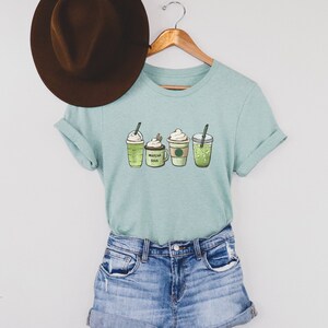 May include: A light blue t-shirt with a graphic of four different green iced coffee drinks. The drinks include a matcha latte, a coffee drink with whipped cream, a green tea latte, and a green iced coffee. The text "MATCHA SOY" is visible on the coffee drink with whipped cream.