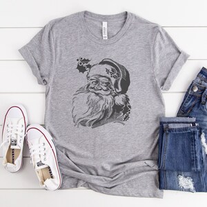 May include: A grey t-shirt featuring a vintage illustration of Santa Claus's face. The illustration is in black and white and shows a detailed drawing of Santa's beard and hat.