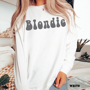 Puede incluir: Camiseta blanca de manga larga con la palabra "Blondie" en gris, con letras de estilo retro en el pecho. La camiseta est&aacute; hecha de un material suave. La palabra "WHITE" est&aacute; impresa en una revista.