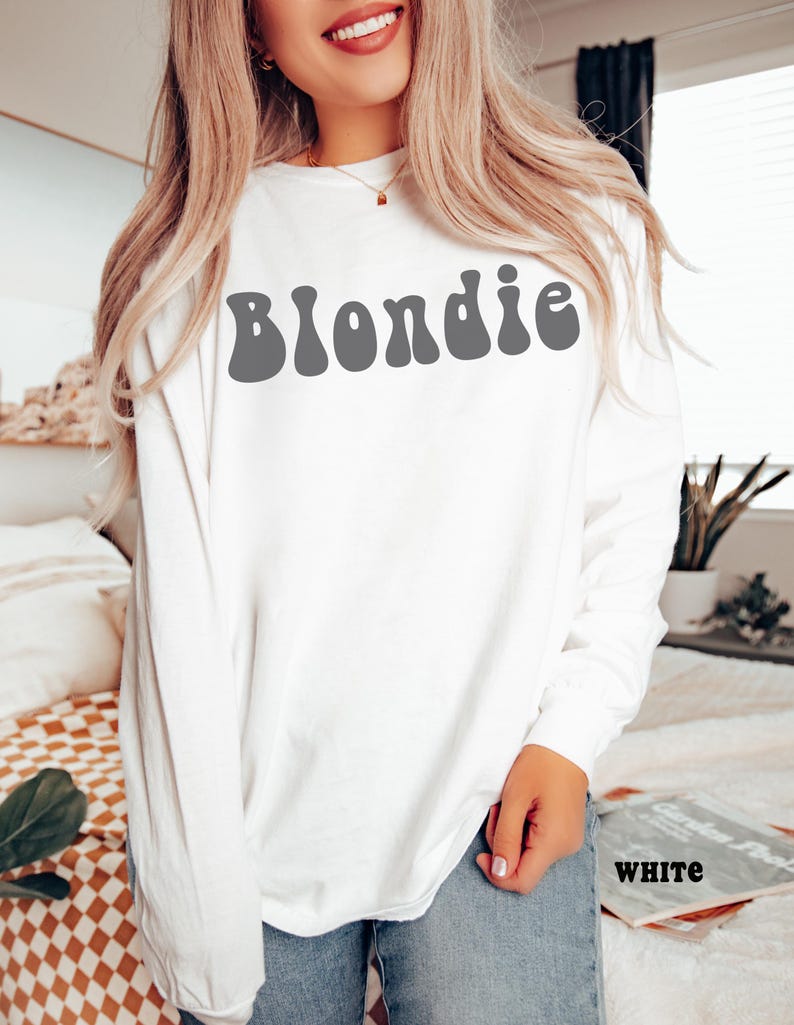 Puede incluir: Camiseta blanca de manga larga con la palabra "Blondie" en gris, con letras de estilo retro en el pecho. La camiseta est&aacute; hecha de un material suave. La palabra "WHITE" est&aacute; impresa en una revista.