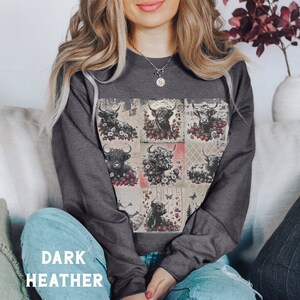 May include: A dark heather grey sweatshirt with a patchwork design featuring black and white Highland cows surrounded by red strawberries and floral accents.