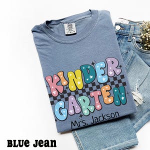 May include: A blue t-shirt with the word "KINDERGARTEN" in colorful bubble letters with stars and a black and white checkered background. The name "Mrs. Jackson" is printed below the word. The shirt is paired with blue jeans.