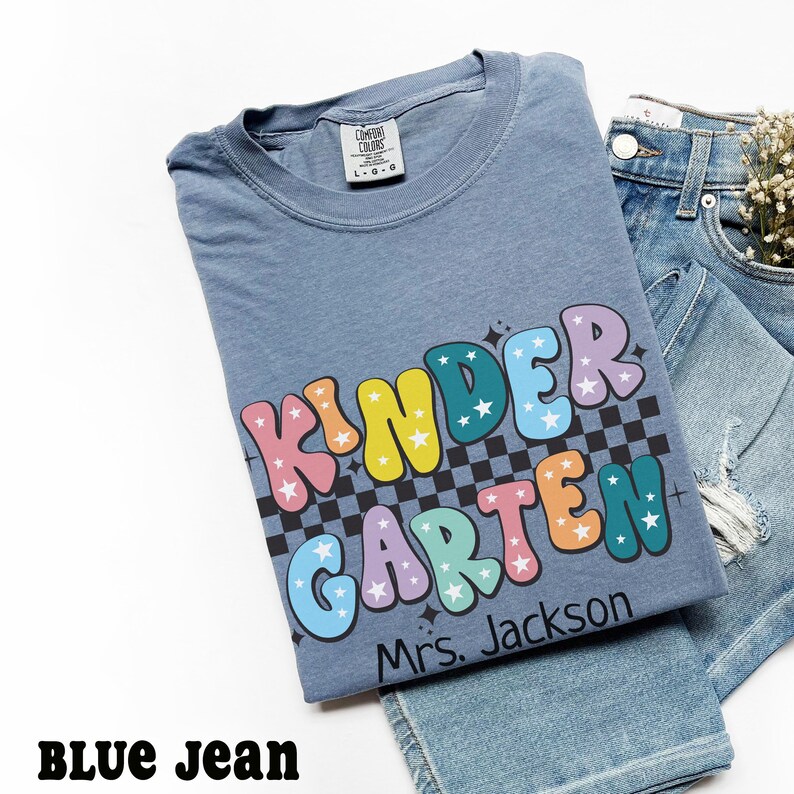 May include: A blue t-shirt with the word "KINDERGARTEN" in colorful bubble letters with stars and a black and white checkered background. The name "Mrs. Jackson" is printed below the word. The shirt is paired with blue jeans.