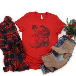 May include: Red t-shirt featuring a vintage illustration of Santa Claus's face. The illustration is in black and white and includes a holly sprig.