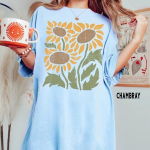May include: A light blue oversized t-shirt with a graphic print of three stylized sunflowers in shades of yellow, brown, and green. The text "CHAMBRAY" is printed in black at the bottom right corner of the shirt.