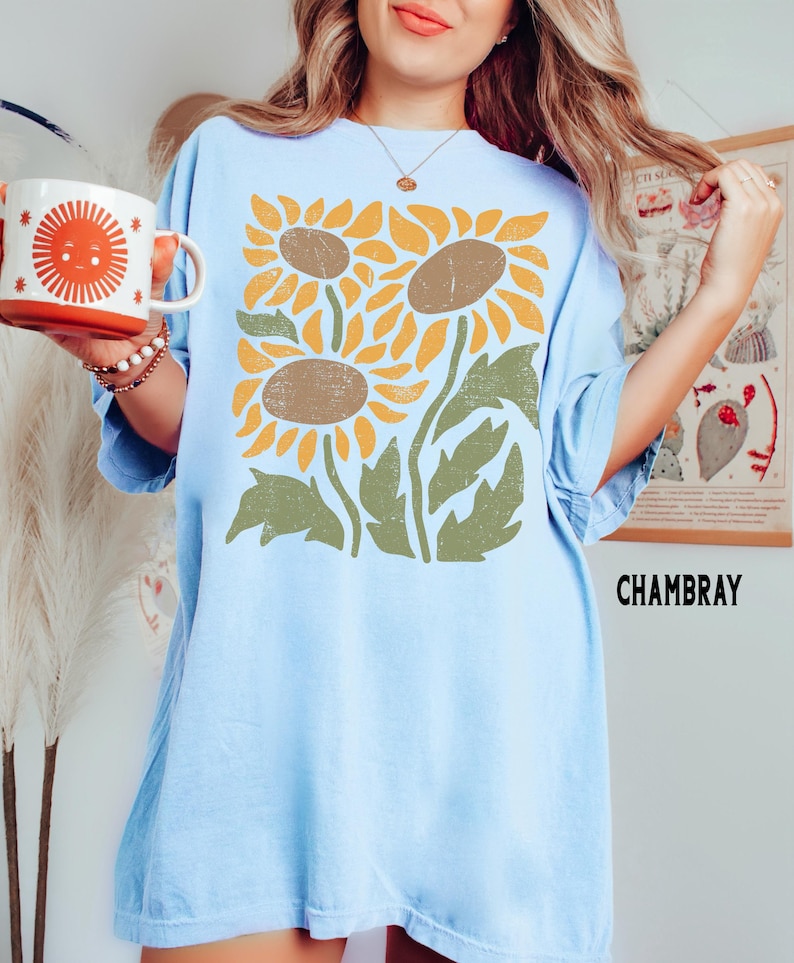 May include: A light blue oversized t-shirt with a graphic print of three stylized sunflowers in shades of yellow, brown, and green. The text "CHAMBRAY" is printed in black at the bottom right corner of the shirt.