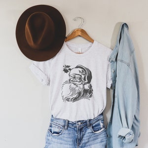 May include: A white t-shirt featuring a vintage illustration of Santa Claus's face in grey. The shirt is hanging on a wooden hanger with a brown hat and a blue denim jacket.