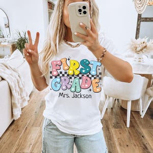 May include: White t-shirt with the words "FIRST GRADE" in colorful, retro-style lettering, over a black and white checkered pattern. The name "Mrs. Jackson" is printed below. The shirt is worn with light blue jeans.