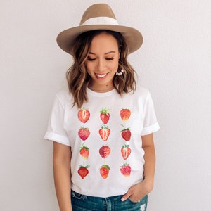Op de afbeelding: Een wit T-shirt met een patroon van rode aardbeien. De aardbeien zijn gerangschikt in een rasterpatroon en zijn allemaal van verschillende grootte en vorm. Het shirt wordt gedragen door een persoon met bruin haar en een bruine hoed.