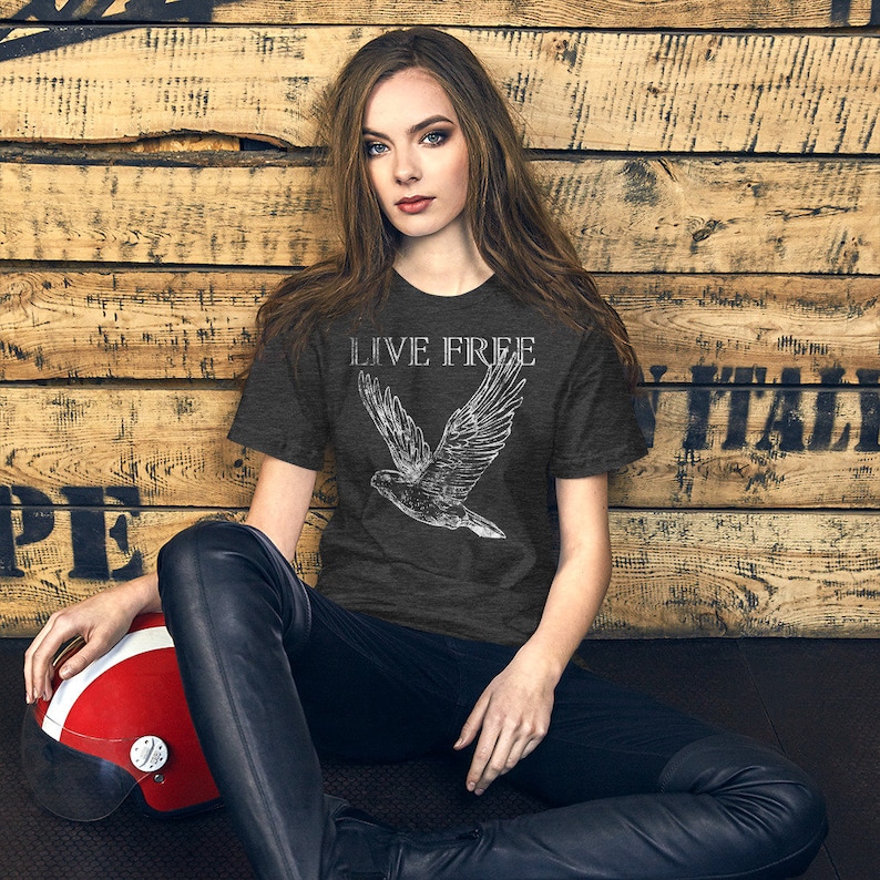 Op de afbeelding: Een vrouw draagt een donkergrijs T-shirt met een witte afbeelding van een vogel in de vlucht en de tekst "LIVE FREE". Ze zit op een houten oppervlak met een rode helm naast haar.
