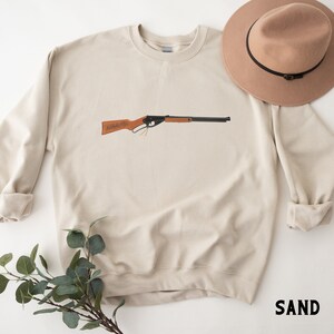 May include: A beige sweatshirt with a brown and black toy rifle printed on the front. The sweatshirt has a crew neck and long sleeves. The word "SAND" is printed in black on the bottom right corner of the image.