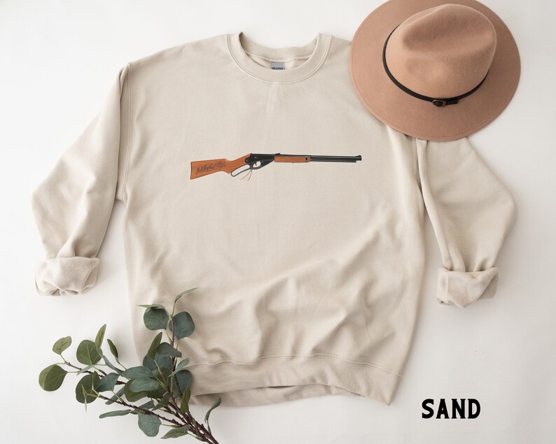 May include: A beige sweatshirt with a brown and black toy rifle printed on the front. The sweatshirt has a crew neck and long sleeves. The word "SAND" is printed in black on the bottom right corner of the image.