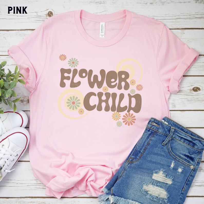 Puede incluir: Camiseta rosa claro con mangas enrolladas, con la frase "FLOWER CHILD" en marr&oacute;n, adornada con motivos florales y espirales. La camiseta se combina con pantalones cortos de mezclilla y zapatillas blancas.