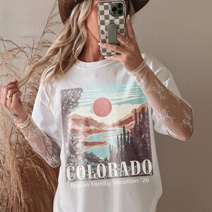 May include: White t-shirt with a Colorado landscape graphic featuring a lake, mountains, and a red sun. The text "COLORADO" and "Brown Family Vacation '26" are printed below the image. The shirt has lace sleeves and is paired with a brown cowboy hat.
