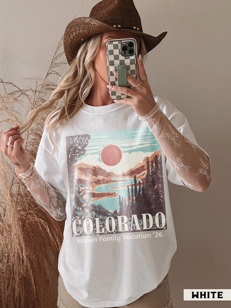 May include: White t-shirt with a Colorado landscape graphic featuring a lake, mountains, and a red sun. The text "COLORADO" and "Brown Family Vacation '26" are printed below the image. The shirt has lace sleeves and is paired with a brown cowboy hat.