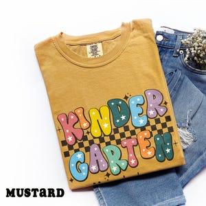 May include: Mustard-colored t-shirt with the word "KINDERGARTEN" in colorful bubble letters with star accents. The shirt is folded and placed on top of a pair of blue jeans.