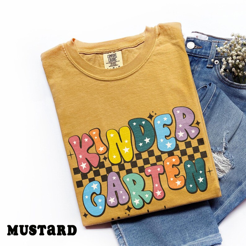 May include: Mustard-colored t-shirt with the word "KINDERGARTEN" in colorful bubble letters with star accents. The shirt is folded and placed on top of a pair of blue jeans.