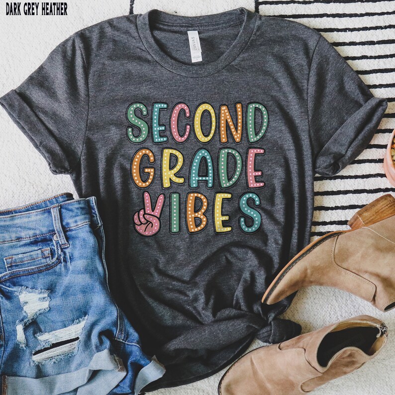 May include: A dark gray heather t-shirt with the text "Second Grade Vibes" in colorful letters with white polka dots. A peace sign is in front of the text.