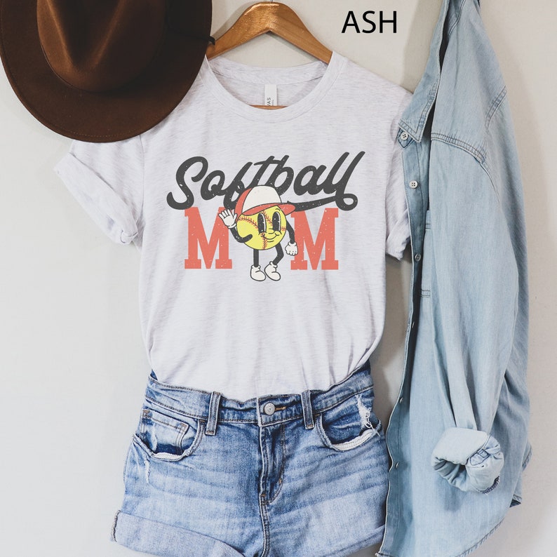 May include: A light gray t-shirt with the words "Softball Mom" in black and red lettering. The shirt features a cartoon baseball character wearing a hat and waving. The shirt is paired with denim shorts and a denim shirt.