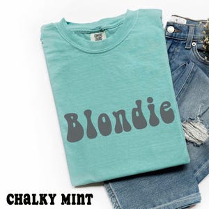 Puede incluir: Una camiseta verde menta con la palabra "Blondie" en letras grises de estilo retro. La camiseta est&aacute; doblada junto a unos vaqueros azules. El texto "Chalky Mint" est&aacute; en la parte inferior de la imagen.