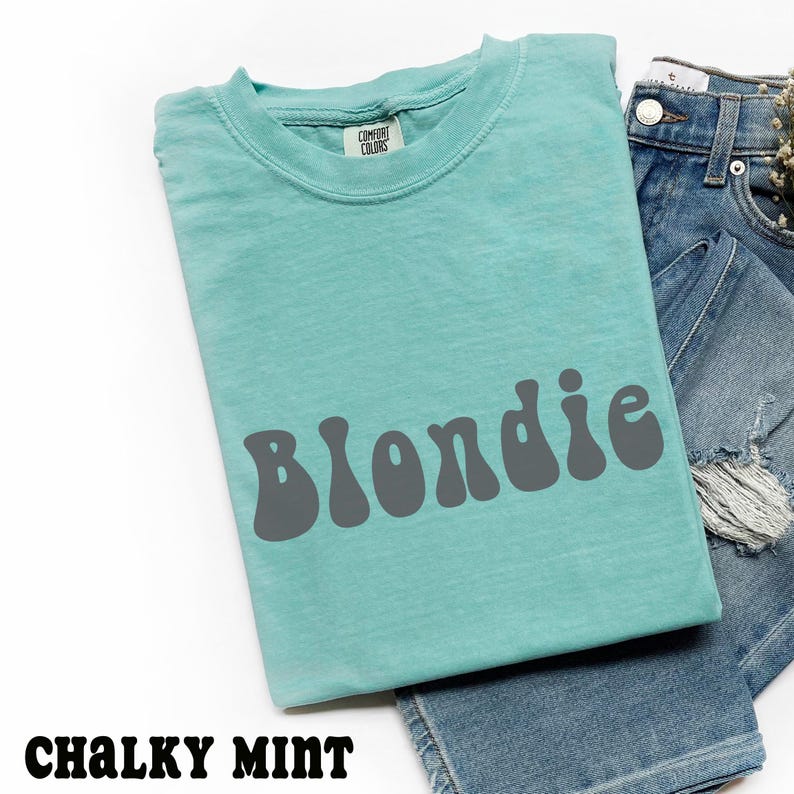 Puede incluir: Una camiseta verde menta con la palabra "Blondie" en letras grises de estilo retro. La camiseta est&aacute; doblada junto a unos vaqueros azules. El texto "Chalky Mint" est&aacute; en la parte inferior de la imagen.