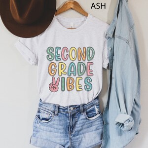 May include: A white t-shirt with the text "SECOND GRADE VIBES" in colorful letters with a peace sign. The shirt is being modeled on a person wearing denim shorts and a denim jacket.