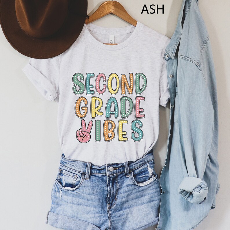 May include: A white t-shirt with the text "SECOND GRADE VIBES" in colorful letters with a peace sign. The shirt is being modeled on a person wearing denim shorts and a denim jacket.