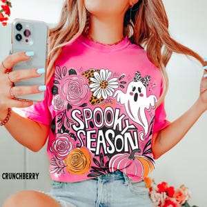 May include: A bright pink t-shirt with a "Spooky Season" graphic. The design includes a white ghost, flowers, and a pumpkin. The word "Crunchberry" is printed at the bottom left.