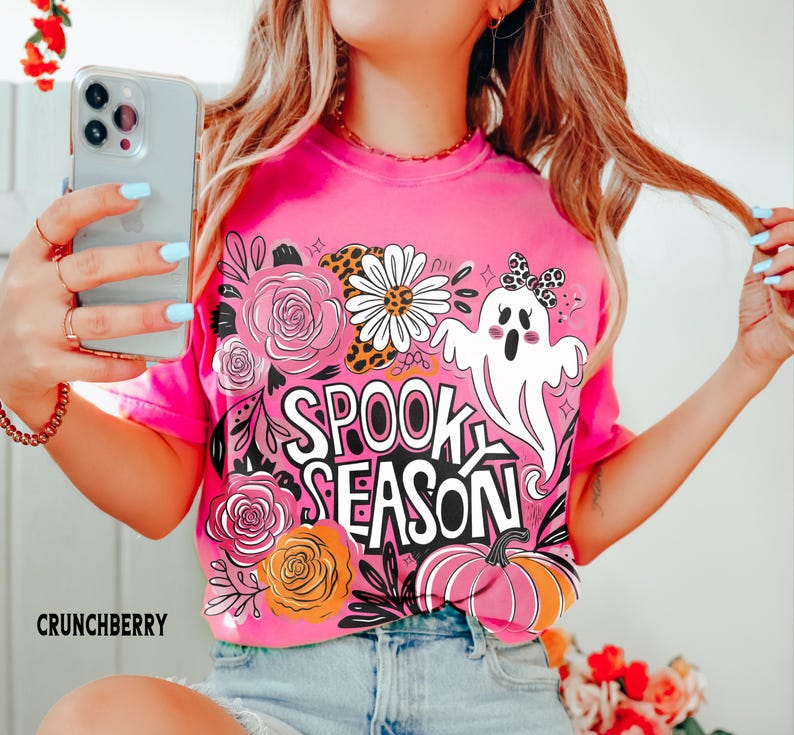 May include: A bright pink t-shirt with a "Spooky Season" graphic. The design includes a white ghost, flowers, and a pumpkin. The word "Crunchberry" is printed at the bottom left.