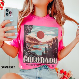 May include: A bright pink t-shirt with a Colorado landscape graphic. The graphic features a lake, mountains, and a sun, with the word "COLORADO" in large letters. The text "Olivia's Bachelorette '26" is also visible.