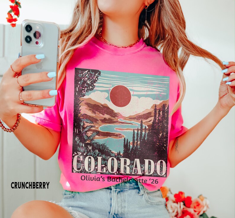 May include: A bright pink t-shirt with a Colorado landscape graphic. The graphic features a lake, mountains, and a sun, with the word "COLORADO" in large letters. The text "Olivia's Bachelorette '26" is also visible.