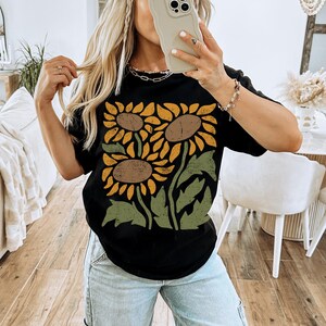 May include: A black t-shirt with a graphic print of three sunflowers in yellow, orange, and brown with green leaves. The text "BLACK" is visible at the bottom of the image.