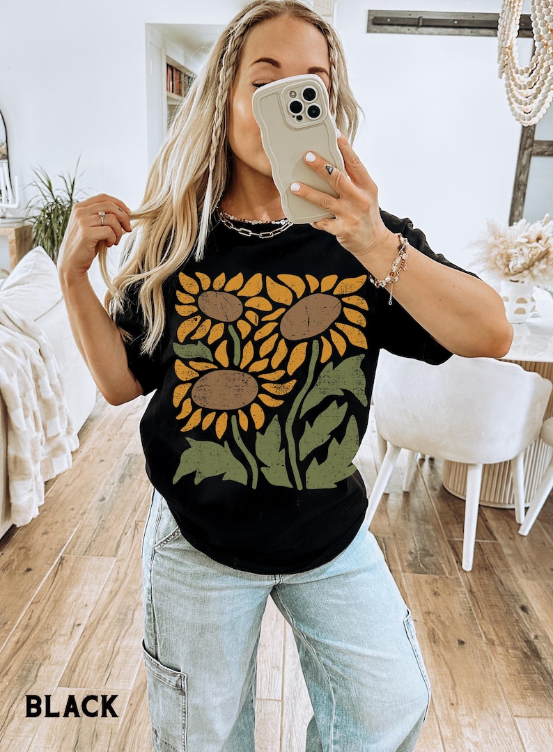 May include: A black t-shirt with a graphic print of three sunflowers in yellow, orange, and brown with green leaves. The text "BLACK" is visible at the bottom of the image.
